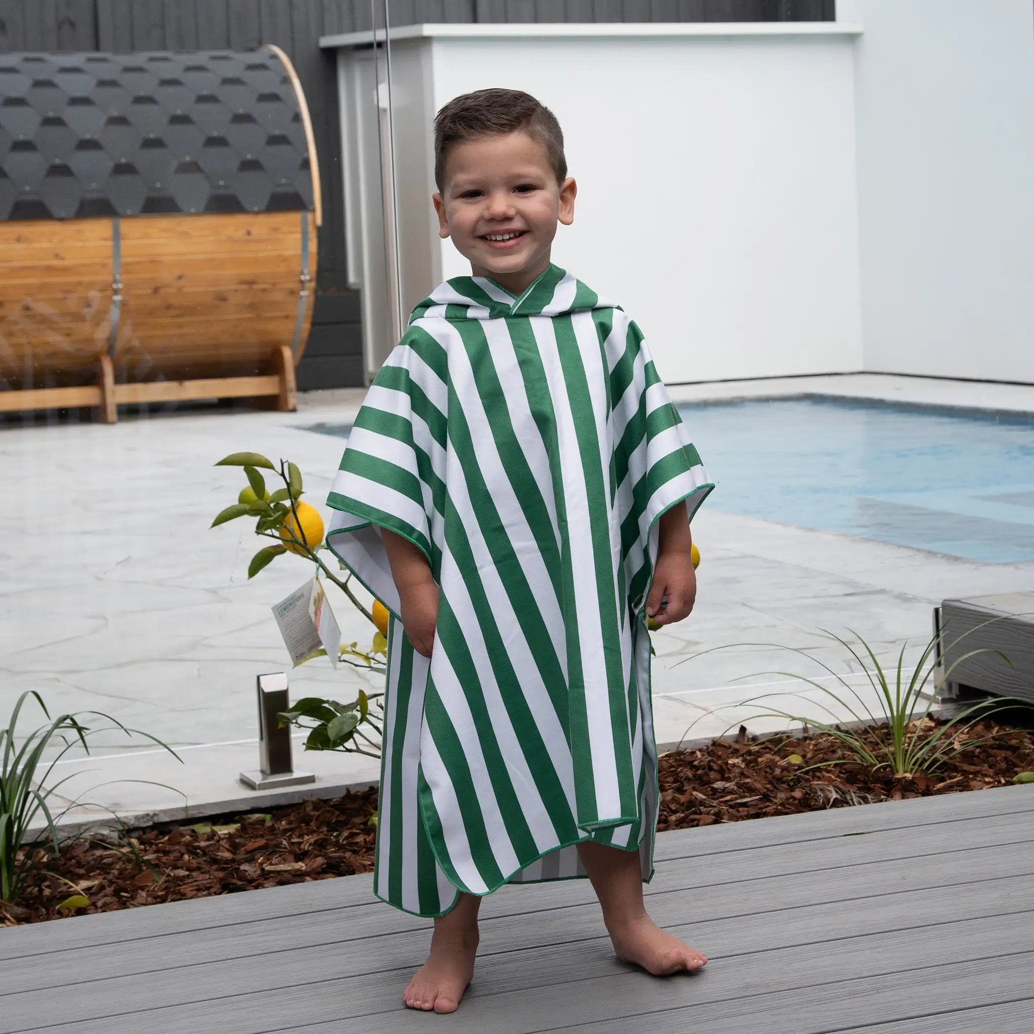 Smiling child wearing a green and white striped StayDry microfibre poncho towel by the pool. Quick-drying, lightweight, and ultra-absorbent for beach, swim, and bath time.